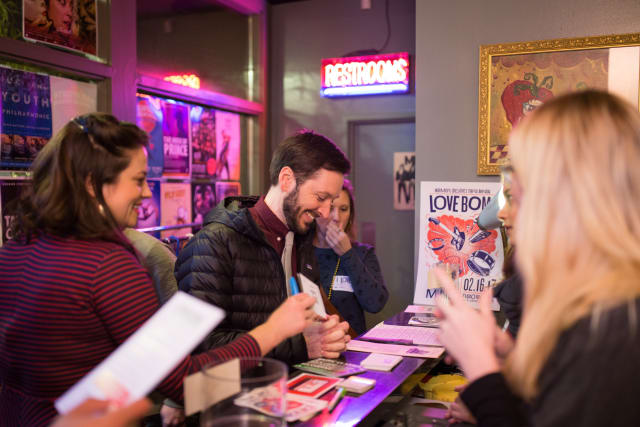 People engaging at the ticket counter during the Love Bomb 2017 event, with colorful neon lights and promotional materials for My Voice Music visible in the background.